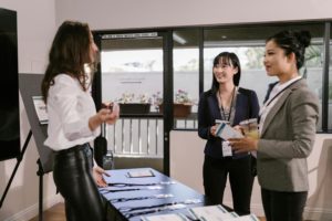 A woman at a registration desk welcomes two professionally dressed attendees during a business event.
