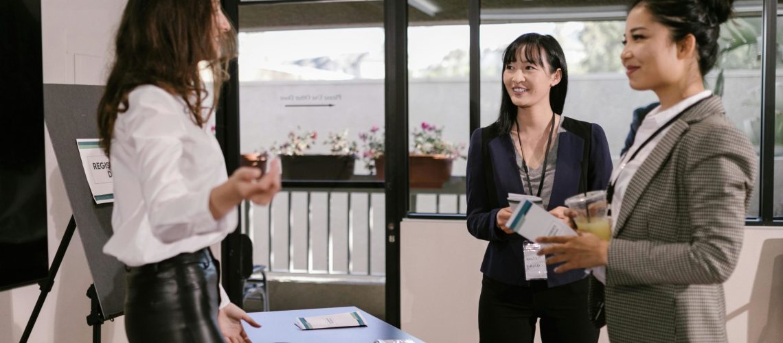 A woman at a registration desk welcomes two professionally dressed attendees during a business event.